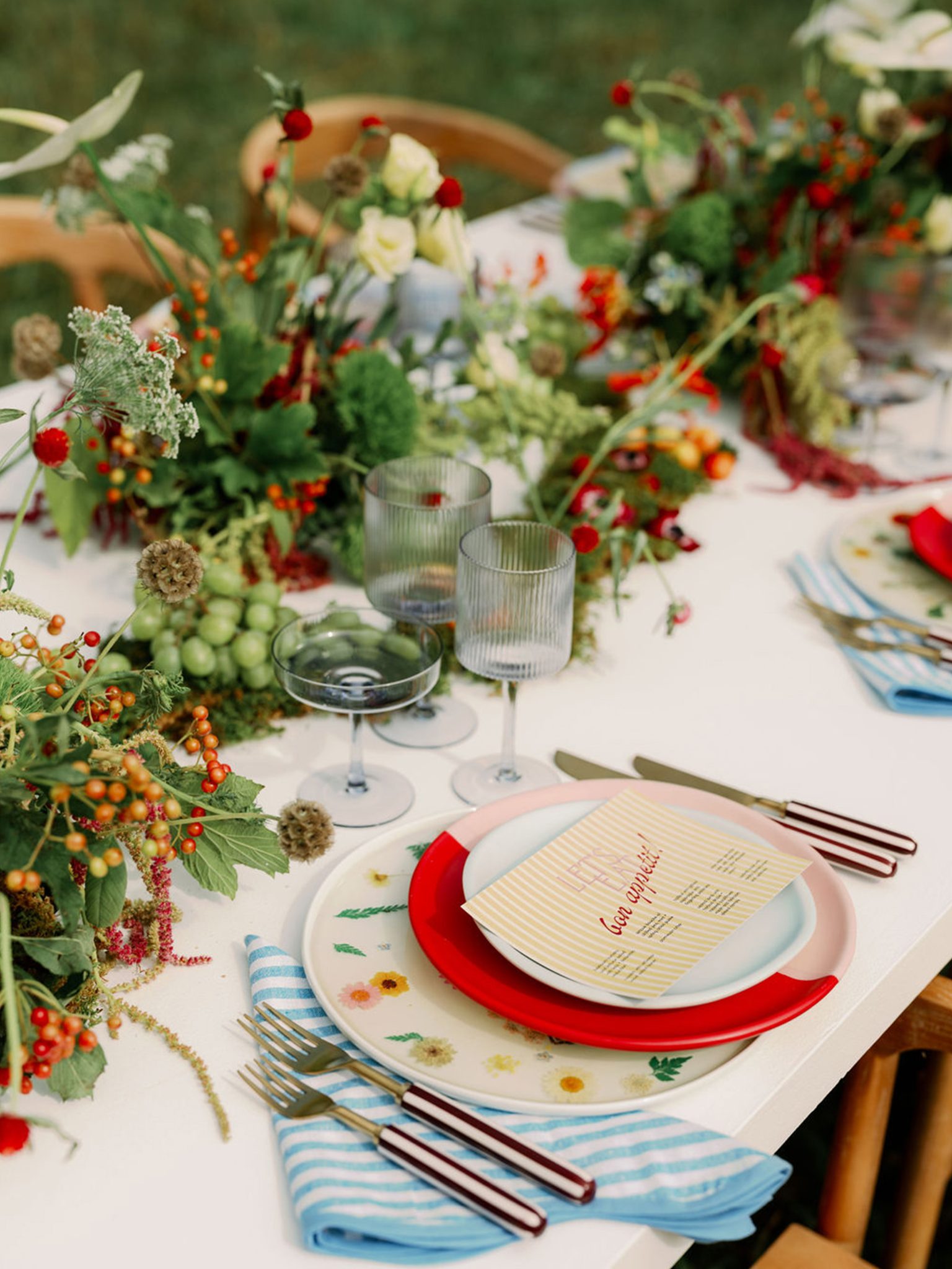 An outdoor tablescape featuring white table tops with lush florals, colorful pressed floral chargers, plates and striped custom menus.