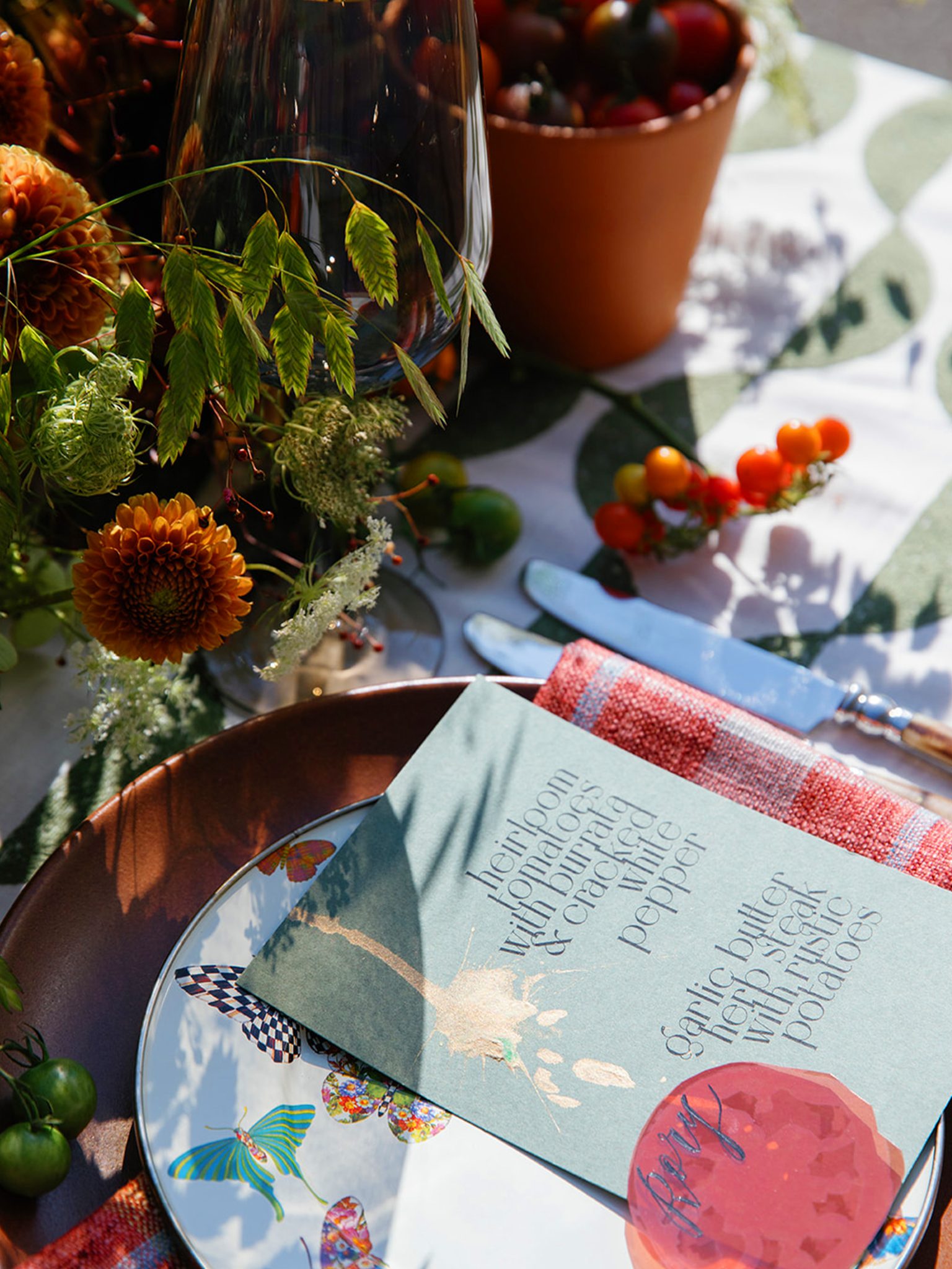 A closeup of a sophisticated tablescape with colorful butterfly plates, green and white linens, heavy modern florals, menus with gold splattered ink and translucent tomato slice place cards.